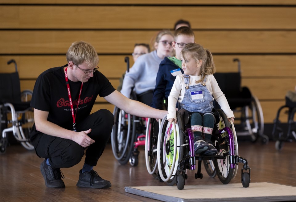 A man in a black t-shirt helps a young girl in a wheelchair to navigate a kerb. Other children in wheelchairs are behind them.
