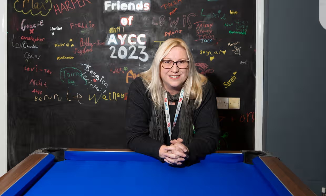 A person leans forward with hands clasped on the edge of a blue pool table, wearing a dark top and a lanyard. They are standing in front of a blackboard wall covered in colorful handwritten names and messages.