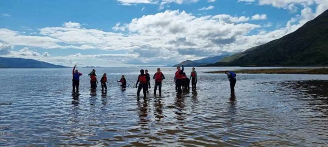 Group of young people standing in shallow coastal water surrounded by hills, taking part in an outdoor activity together.