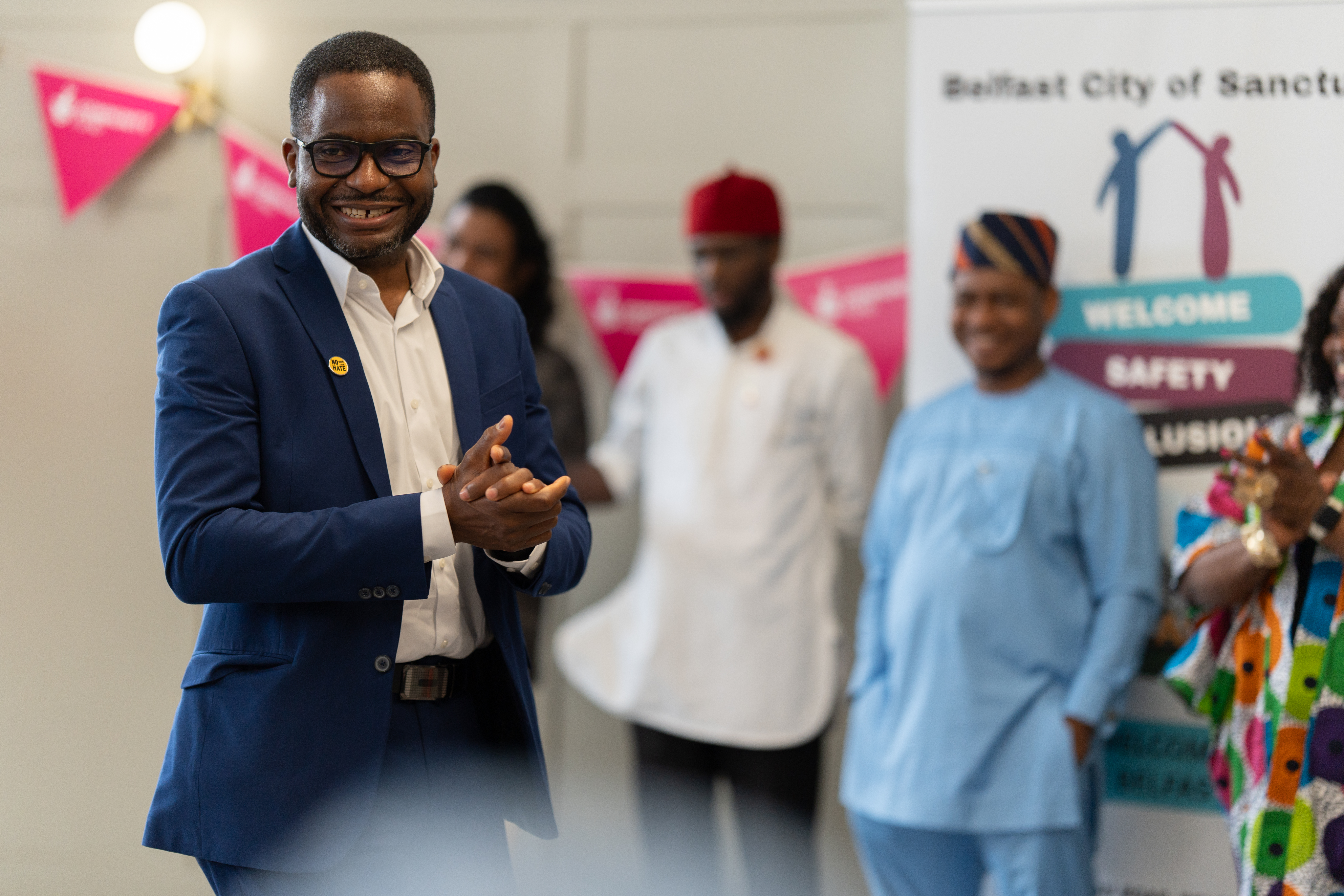 A person smiling wearing a blue suit and white shirt. Out of focus in the background are people lined up smiling at a community event.