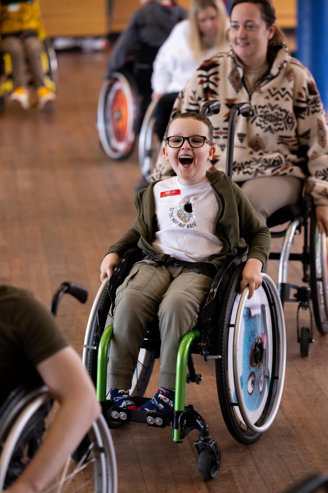 A young boy in a wheelchair smiles at the camera. Other people in wheelchairs are behind him. 