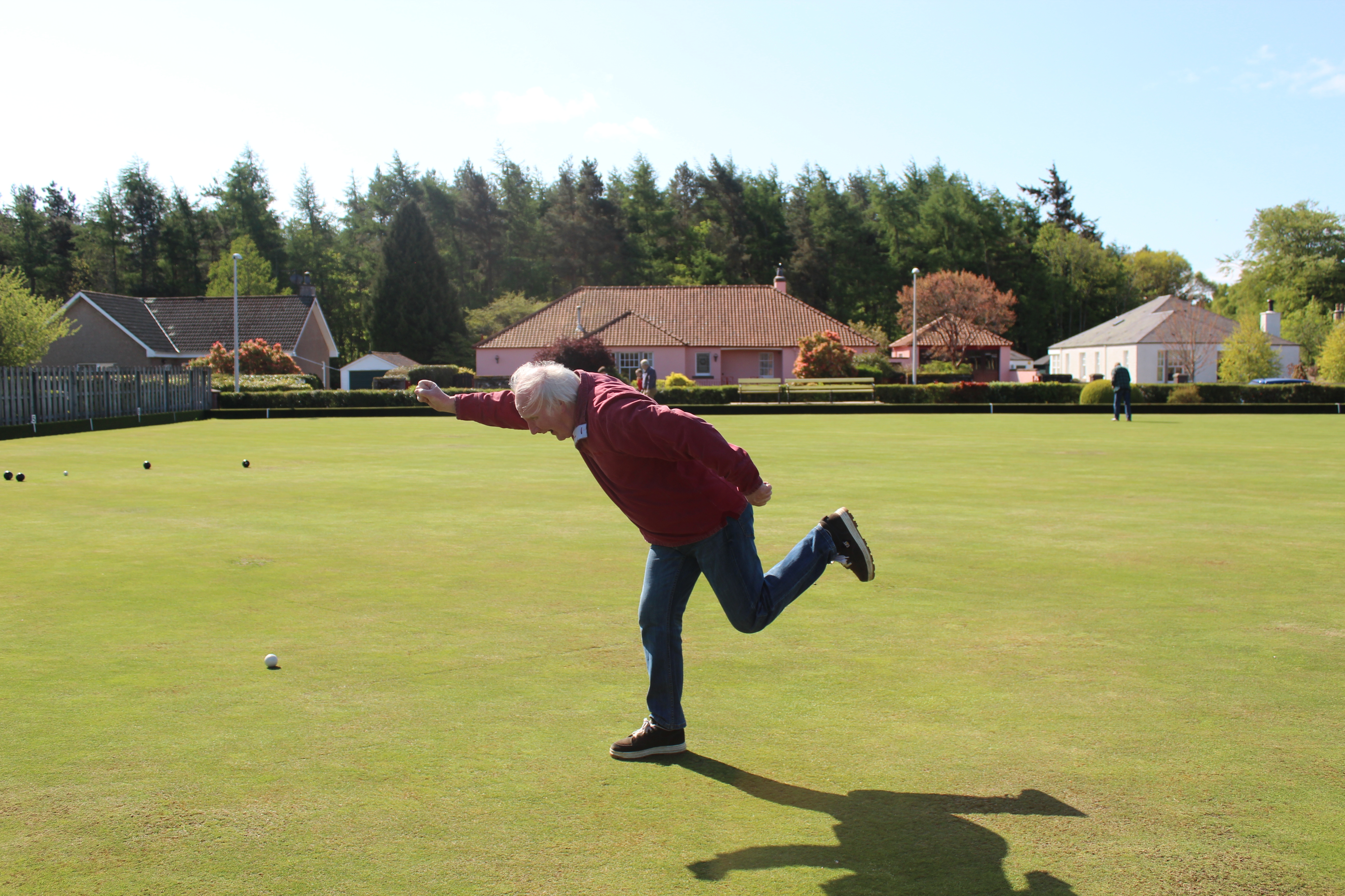 An older man enthusiastically throws a ball on a picturesque bowling green.