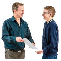 A man hands a paper to a second man. They are both smiling