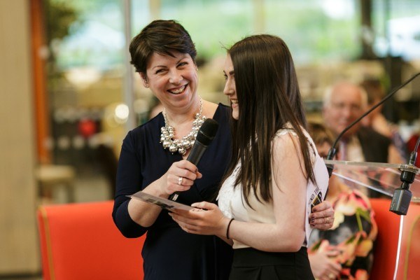 A young woman graduating from WeMindTheGap speaks into a microphone held by Diane Aplin