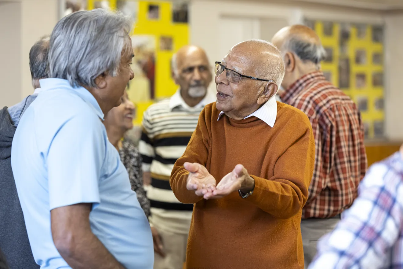 An older man stands among a group of peers. He is speaking to another old man, gesticulating.