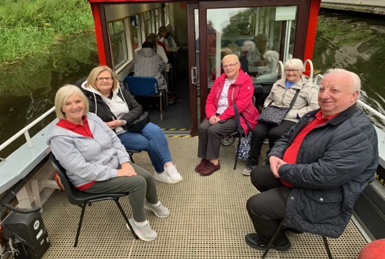 Five smiling members of the Active Seniors project on the open deck area of a small canal boat, with greenery and water visible alongside them.