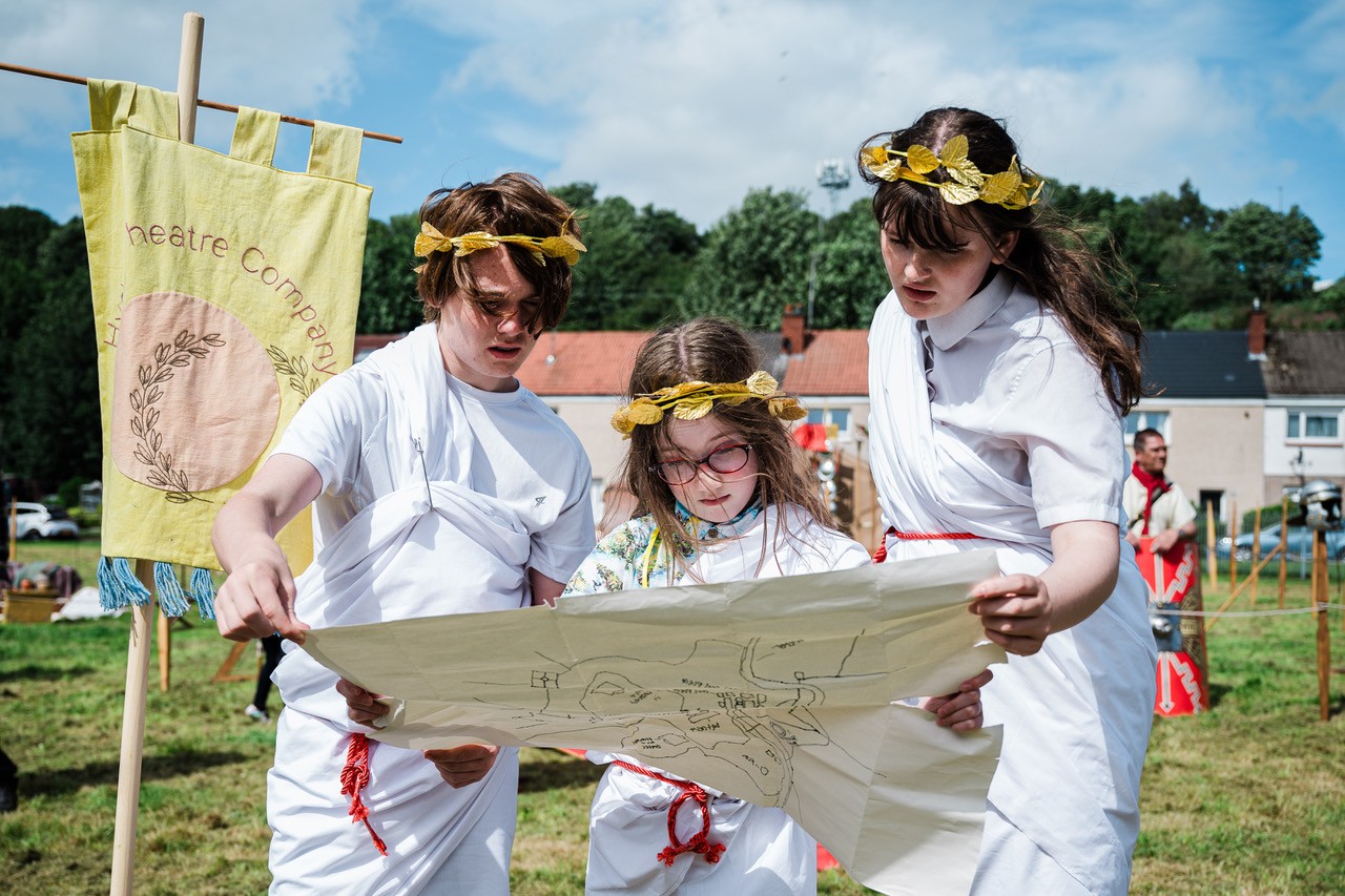 Three young people dressed in toga-style costumes standing in a field reading a hand-drawn map