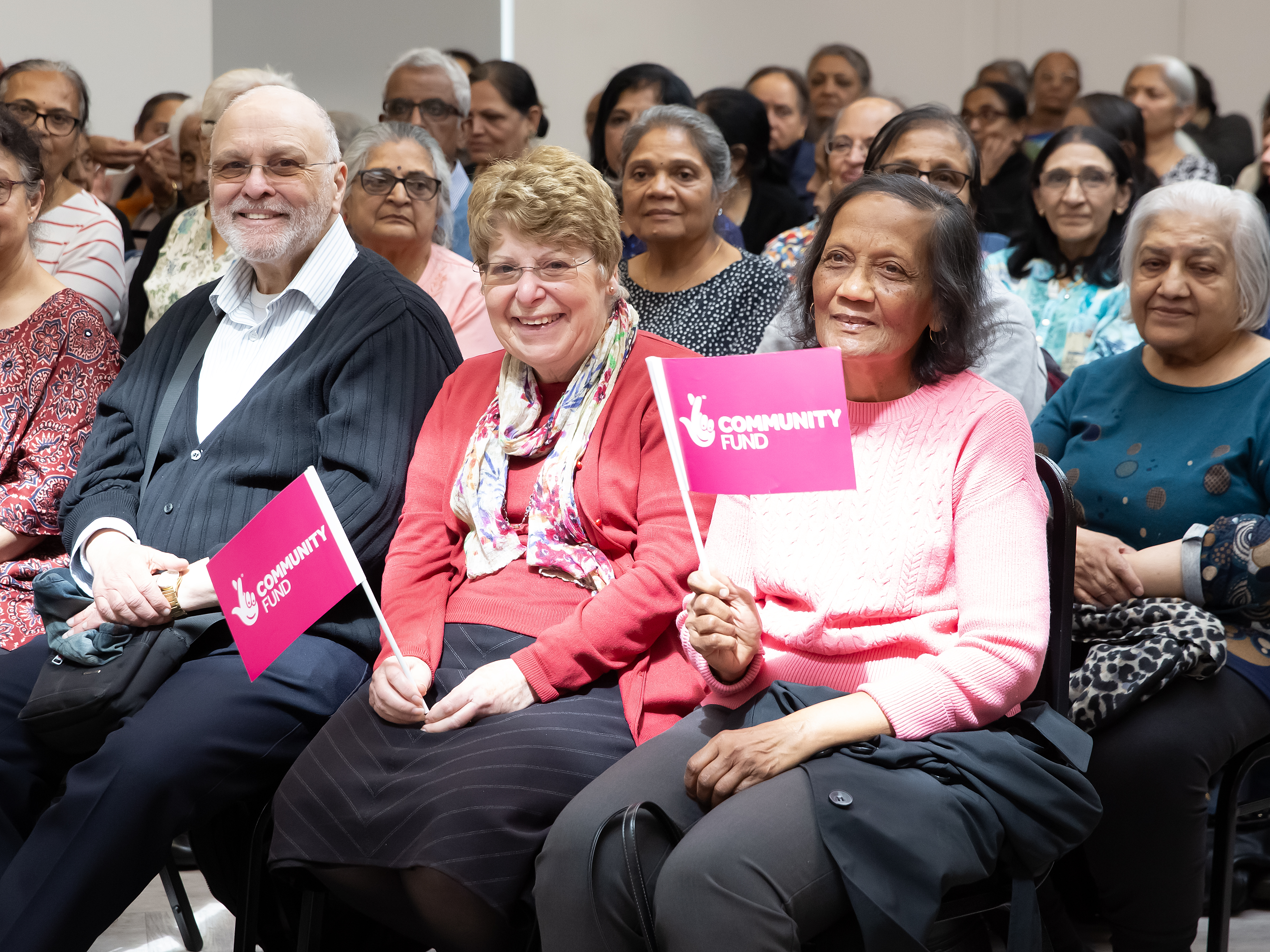 People seated at an event, with a few in the front row holding pink 'community fund' flags.