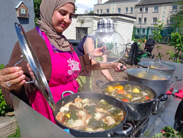 A woman wearing a pink apron and a headscarf lifts the lid from a large pot of food cooking outdoors during a Granton Community Gardeners community event.