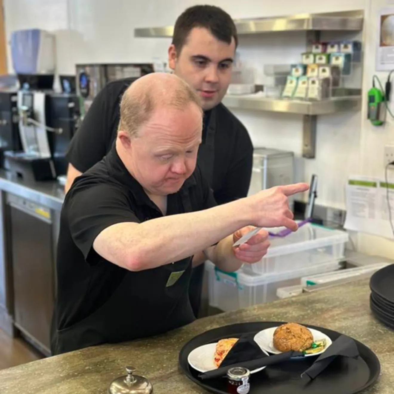 Two people in a kitchen with a plate of food, one person is checking the meal order.