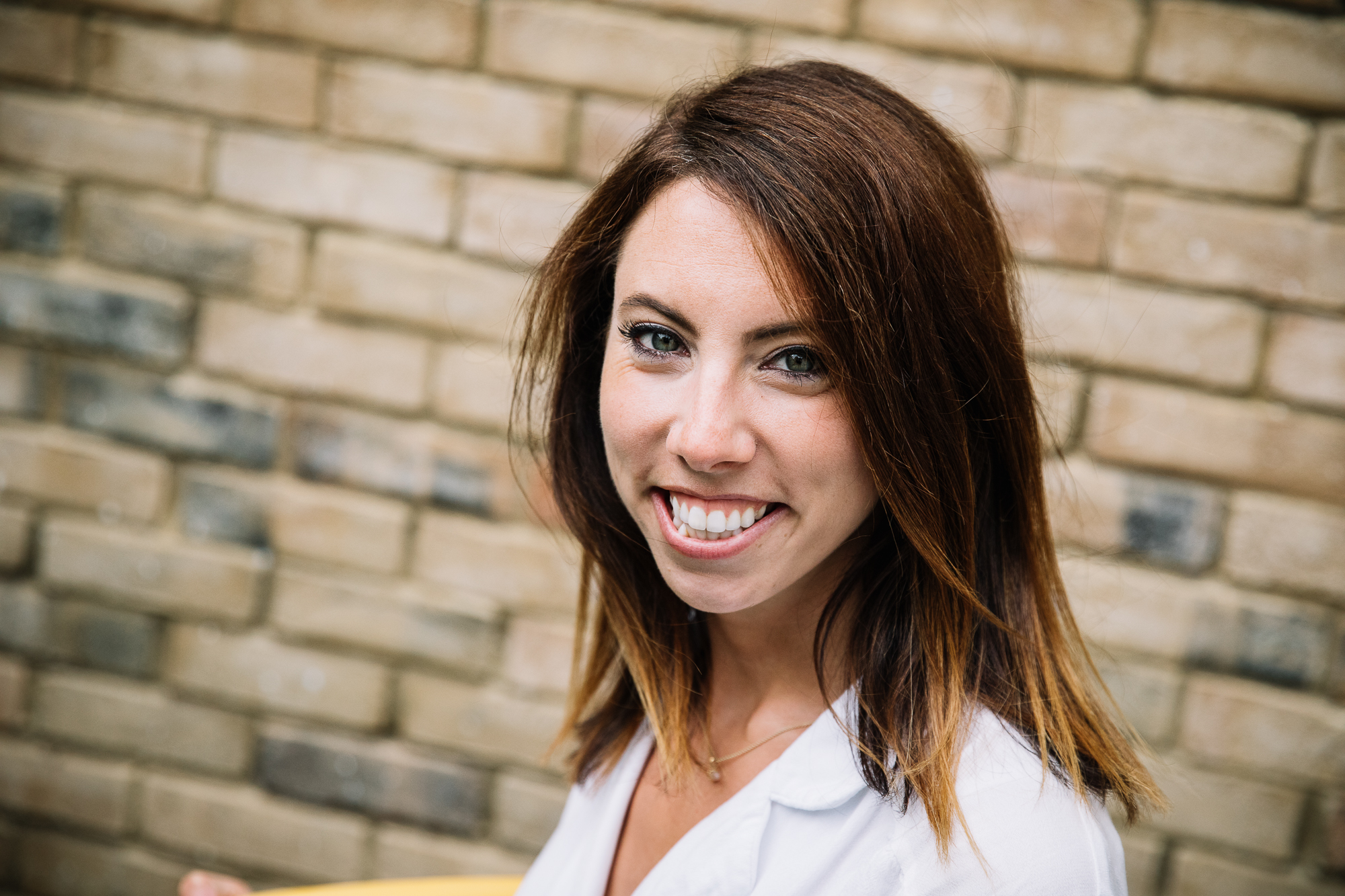 Close-up portrait of a person with shoulder-length brown hair, wearing a top and necklace, standing in front of a blurred brick wall.