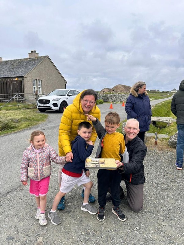 A woman in a yellow jacket and a man kneeling beside her smile at the camera with 3 children on a rural roadside on the Isle of Lewis. They hold a small handmade model on a board from the project.
