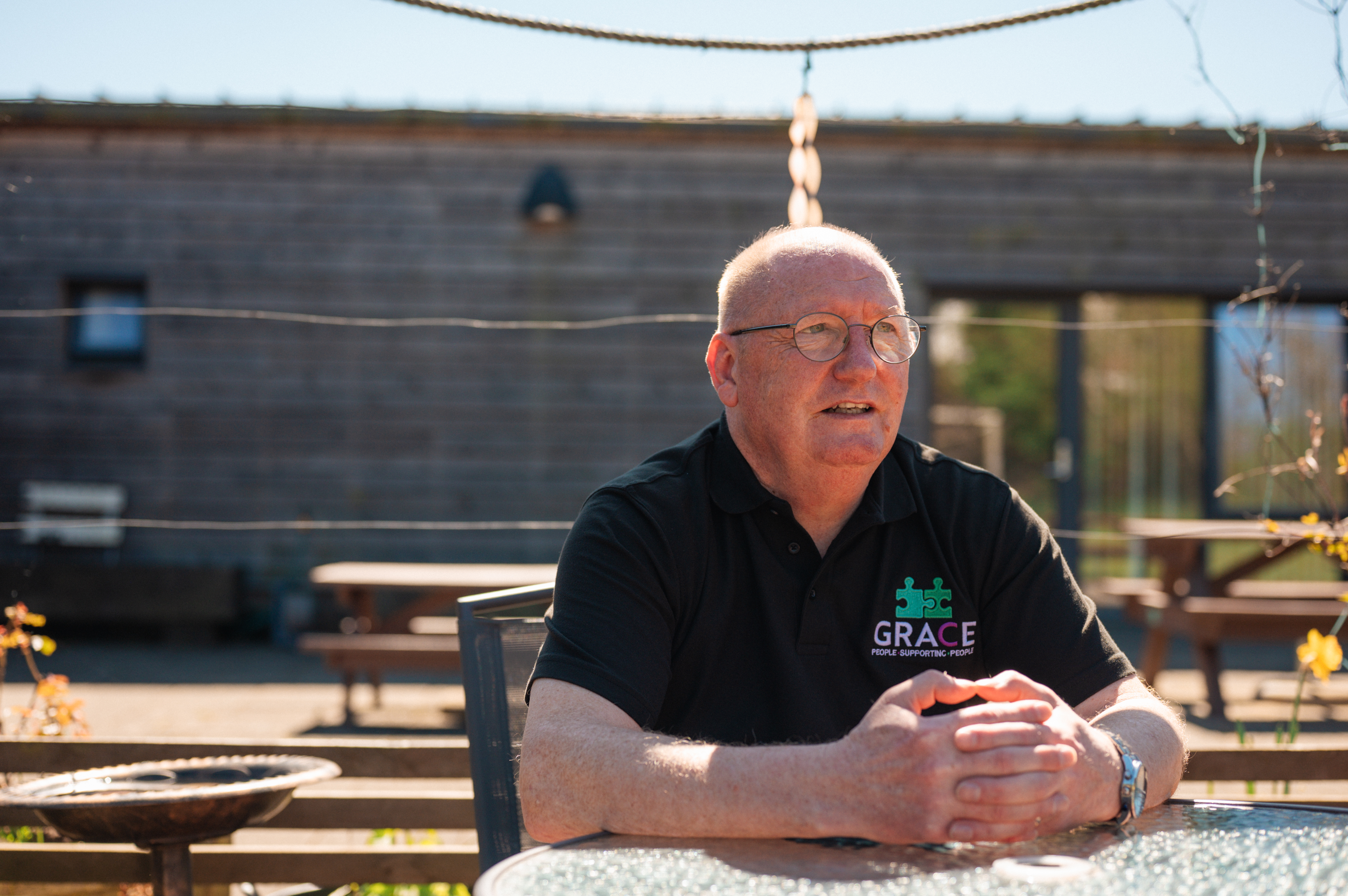 A person wearing glasses leaning on a table outside with their fingers interlocked on the table.
