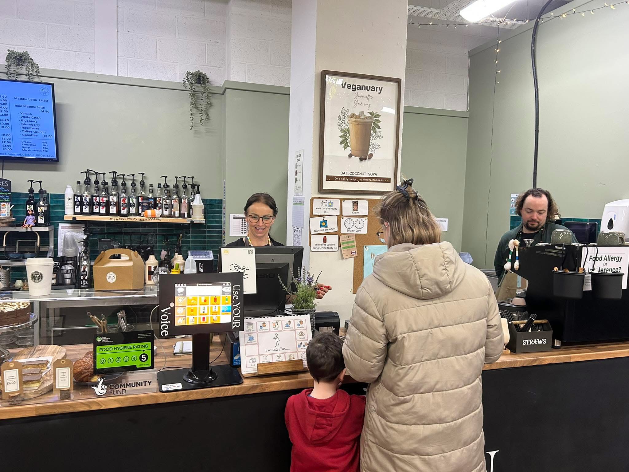 Customer with child ordering drinks at a café counter with menu screen, syrup bottles, and vegan drink poster.