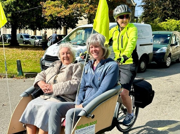 Three people smiling at the camera on a rickshaw bike, with 2 people seated in front and one person pedalling behind.