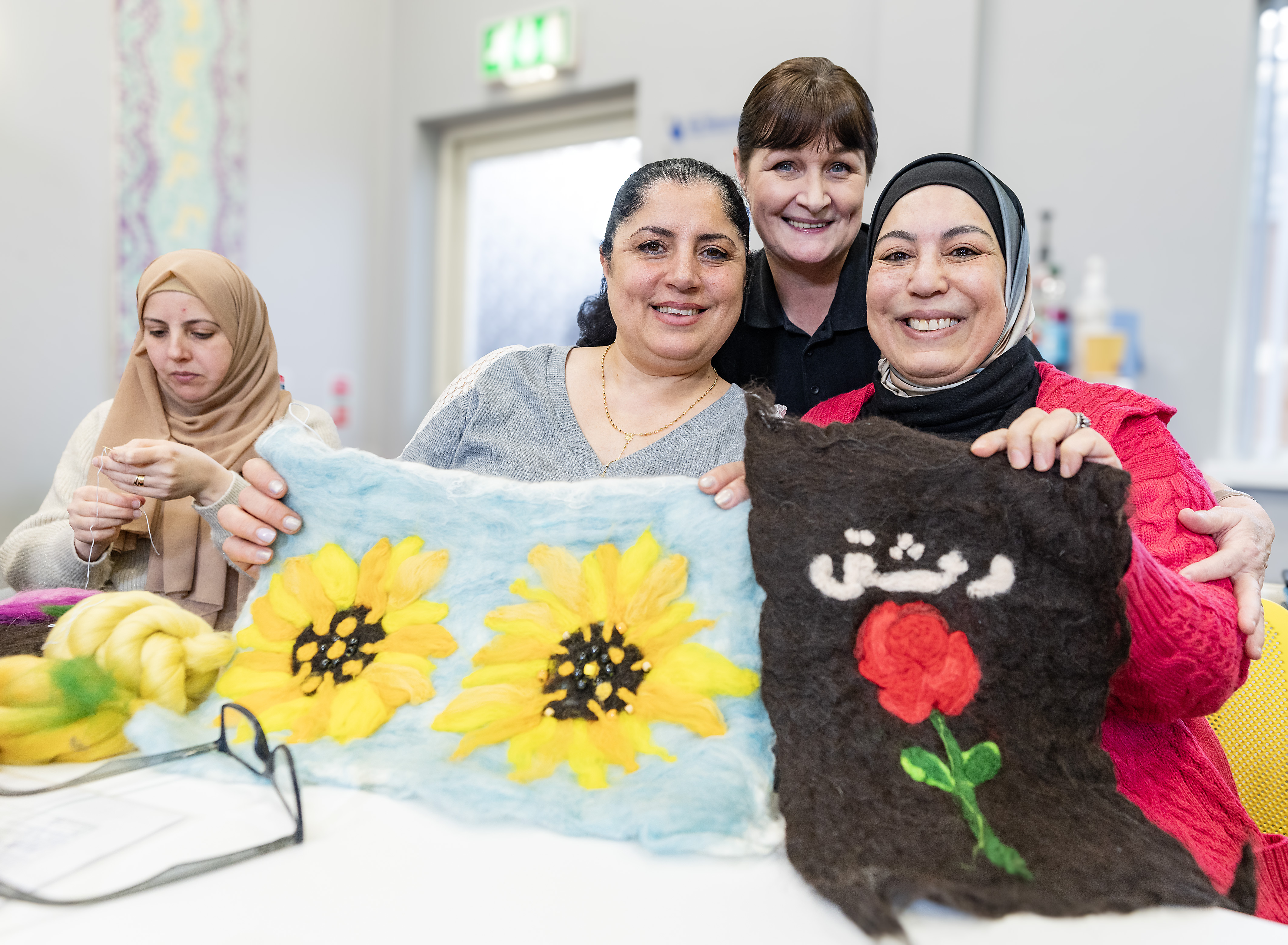 Two women hold up a floral fabric design, smiling. Another woman stands behind them, also smiling. A fourth woman, far left, concentrates on her own design. 