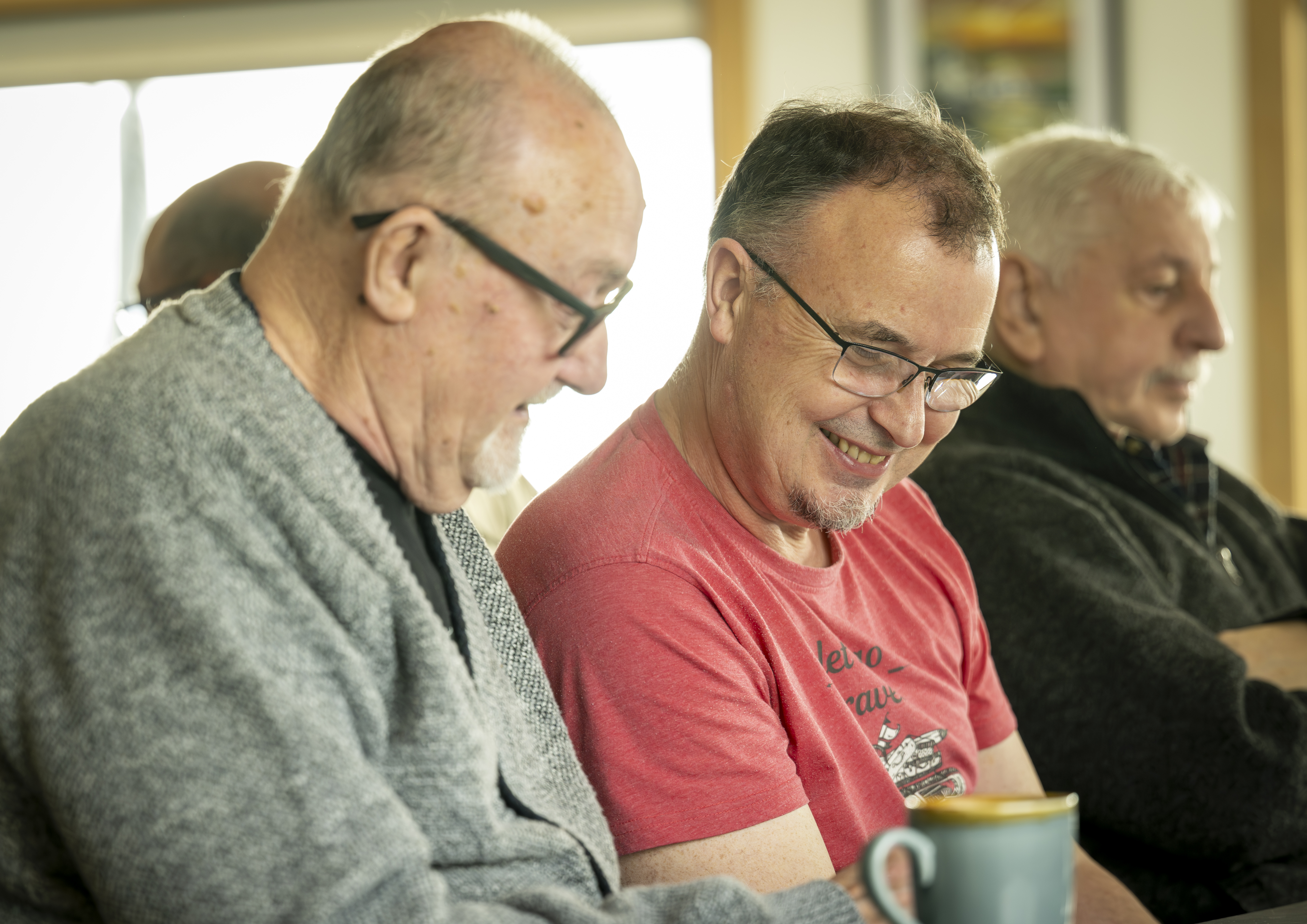 Three adults sitting closely together indoors, two looking down at something in front of them, with a mug visible in the foreground