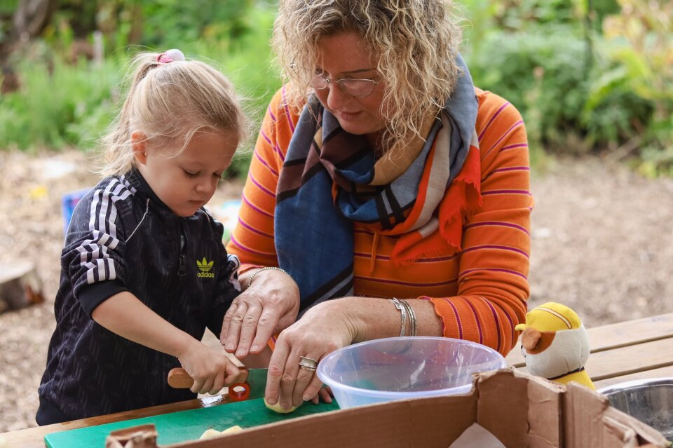 An adult helps a child cut food at an outdoor table.